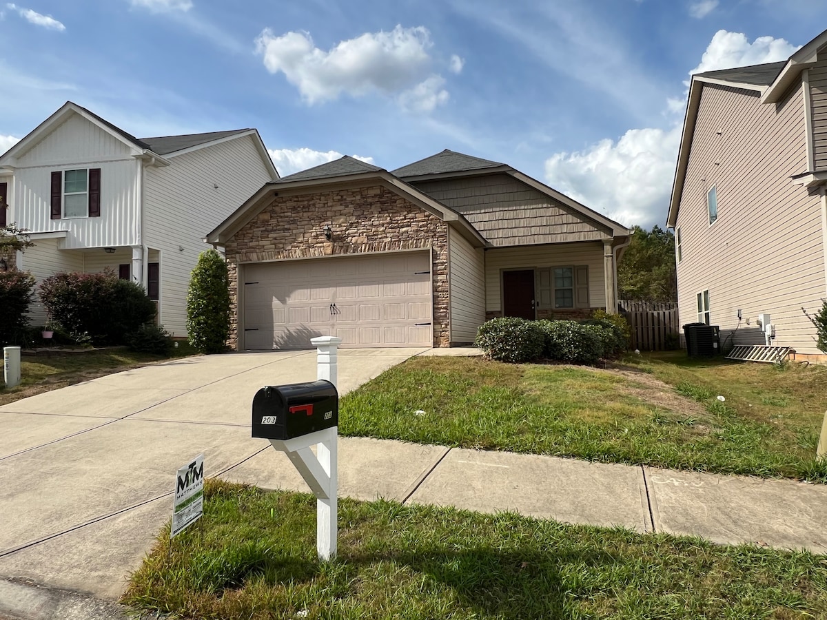 The front exterior of a single-story home is shown, featuring a stone-accented facade and a two-car garage. A neatly kept driveway leads to the entrance, while a well-maintained lawn and shrubs frame the property. Nearby houses are partially visible on either side.