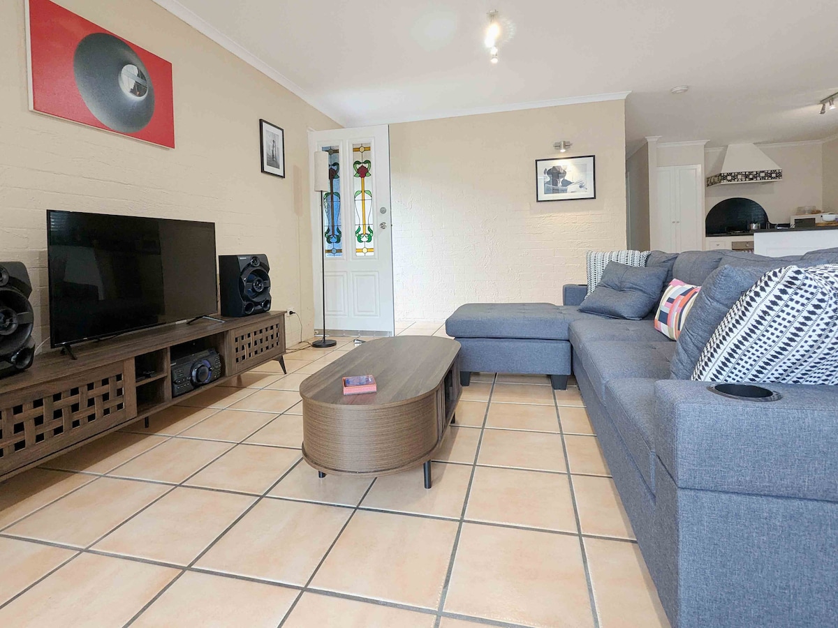 A tiled open-plan living area features a comfortable gray sectional sofa and a low coffee table at its center. A large television is positioned on a media unit, complemented by speakers on either side. Bright wall art adds visual interest, while natural light filters through nearby doors.