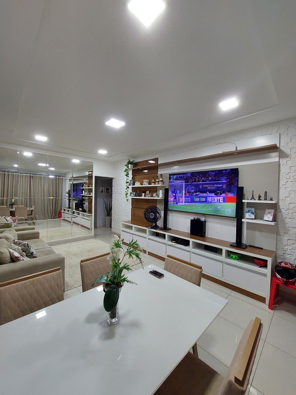 A modern living area features a white entertainment center with a mounted television displaying a sports event. A dining table with neutral-toned chairs sits in the foreground, complemented by a small floral centerpiece. Lighted shelves provide space for decorative items.