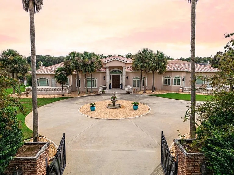 A spacious estate is showcased with a wide driveway leading to the entrance. Lush palm trees frame the house, which is adorned with numerous windows. A central fountain adds a serene element to the landscaped surroundings, creating an inviting entrance.