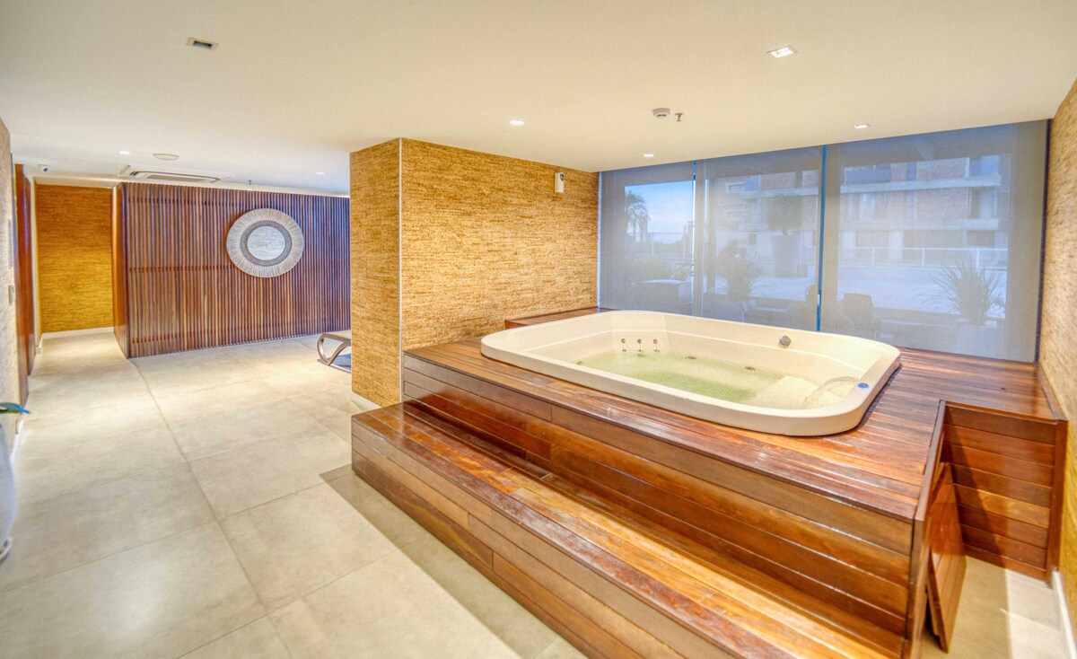 A relaxing area features a wooden platform with a spacious hot tub set against a backdrop of light-colored walls. Natural light filters through sheer panels, creating a soothing environment. The surrounding space includes a smooth concrete floor and minimalistic design elements.