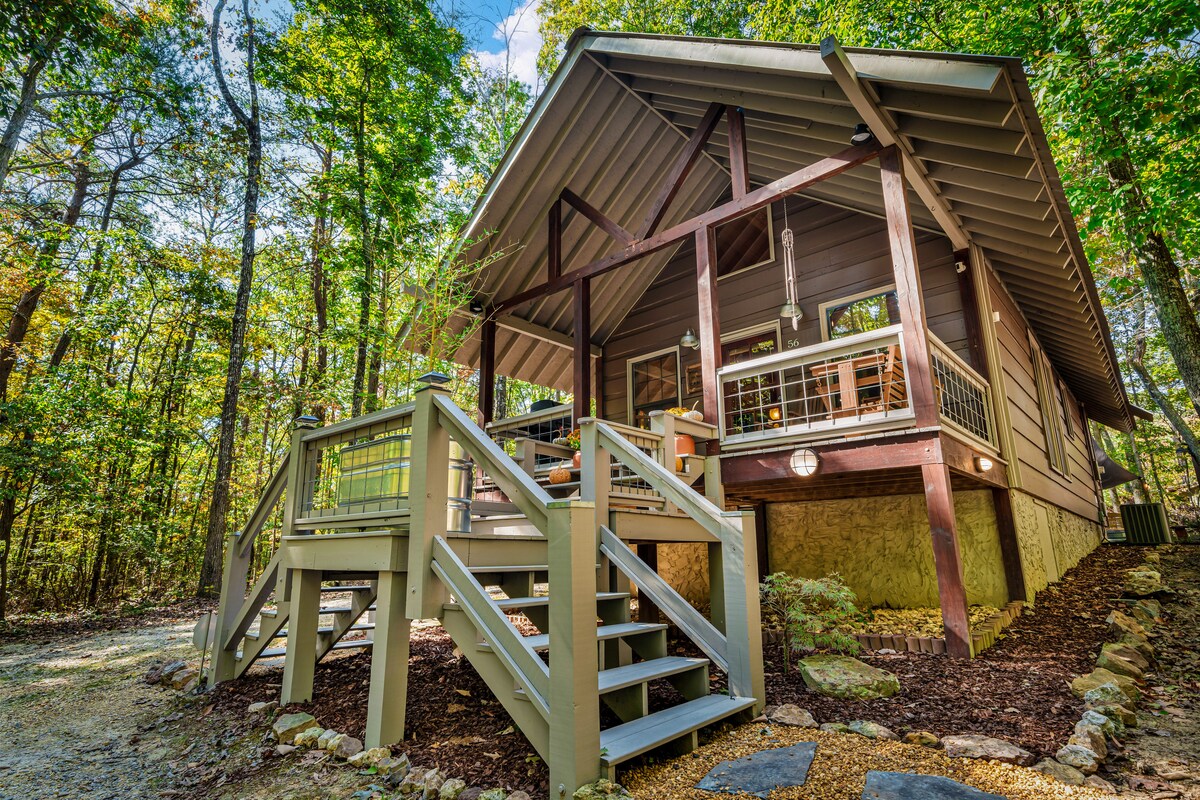 The exterior of a cabin features a welcoming front deck with steps leading up. Surrounded by trees, the structure displays a mix of natural and modern elements, with large windows inviting ample sunlight into the interior.