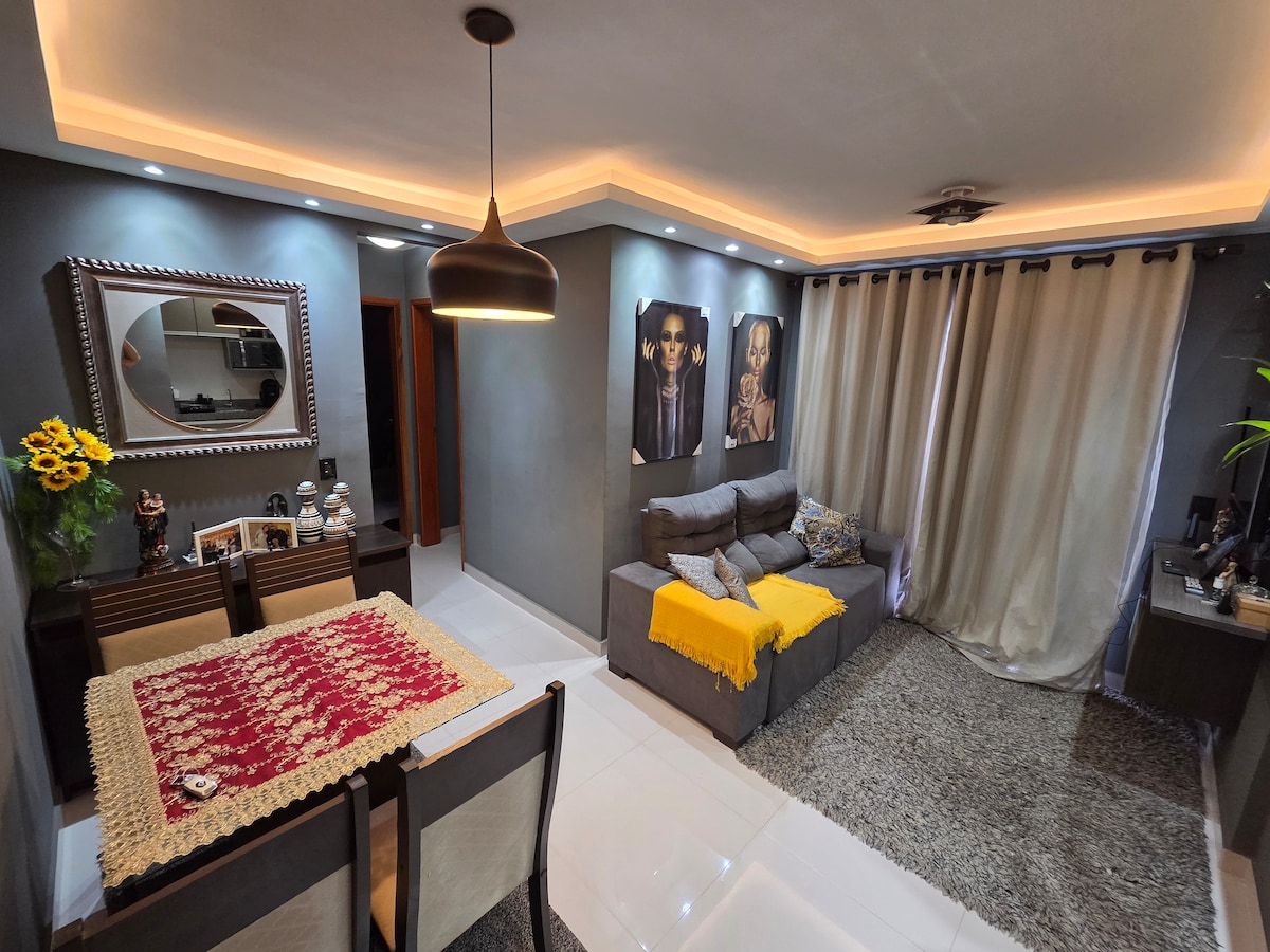A modern living area features a comfortable gray sofa adorned with a yellow throw. A dining table is set with a textured red and cream table runner, while decorative wall art and a framed mirror enhance the space. Soft lighting complements the elegant curtains.