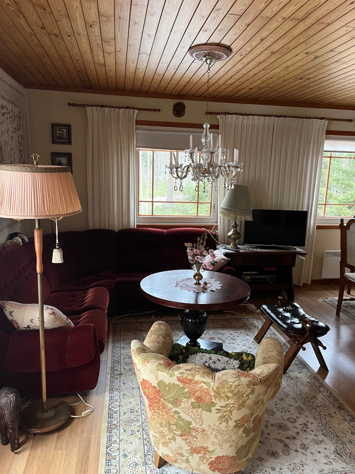 A cozy living area features a velvet red sofa and an elegant chandelier. A round table stands at the center, accompanied by a floral armchair. Natural light filters through large windows, illuminating the hardwood floor and adding warmth to the space.