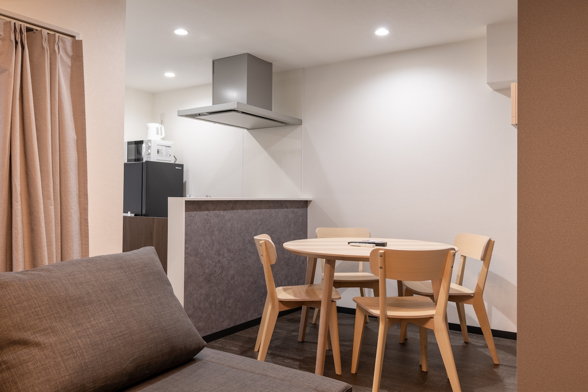 A compact dining area features a round wooden table surrounded by four light-colored chairs. In the background, kitchen appliances are visible, including a microwave and a refrigerator. Soft lighting illuminates the space, enhancing the modern design and functionality.