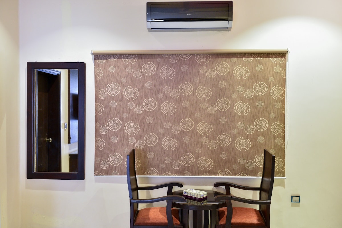 A simple seating area is presented with two dark wooden chairs featuring orange cushions set around a small circular table. Above, a wall-mounted air conditioning unit is visible. A decorative brown roller shade covers the window, and a framed mirror reflects part of the room.