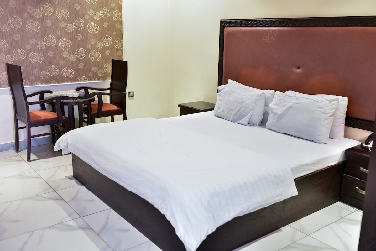 A well-lit sleeping area features a spacious bed with neatly arranged bedding. A small round table with two chairs is positioned nearby, creating a cozy seating arrangement. The walls display a subtle patterned design, enhancing the room's overall ambiance.