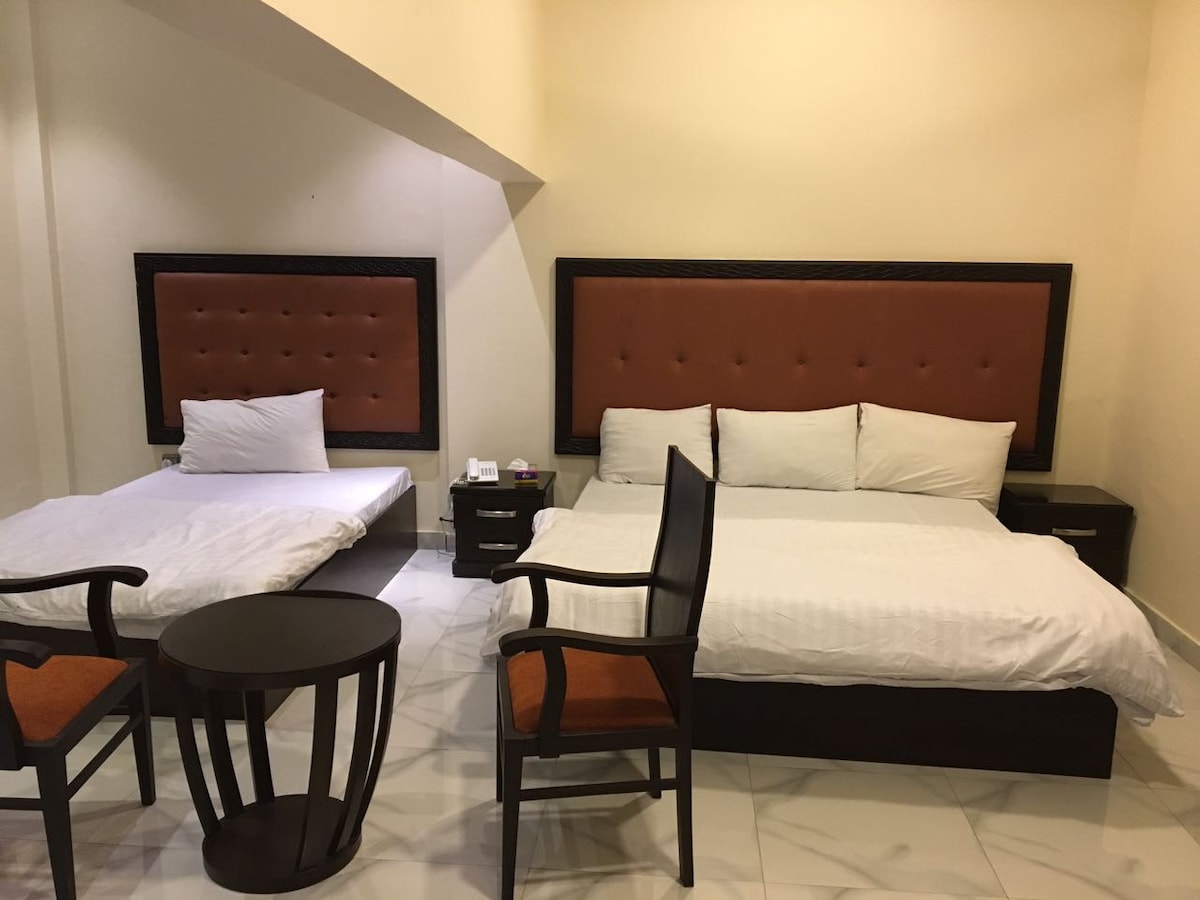 A guest room features two beds with cushioned headboards, one larger and one smaller. A round, dark wood table is positioned between two wooden chairs with orange cushions. The room is brightened by soft overhead lighting and is finished with light-colored tiled flooring.
