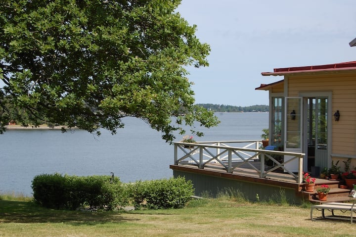 A spacious deck is seen extending from the house, bordered by potted plants and overlooking the calm waters of the lake. A large tree provides shade to the grassy area, enhancing the tranquil setting.