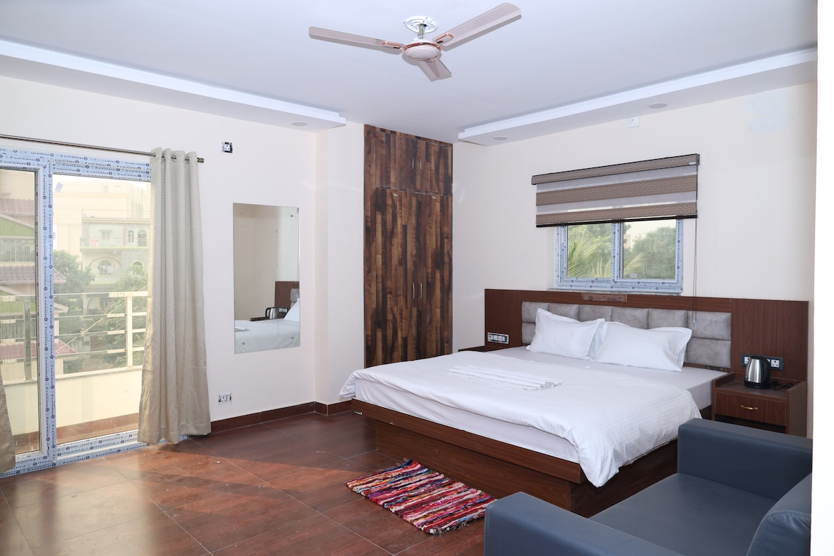 A spacious room is shown with a king size bed and a large window that allows natural light to enter. A dark sofa is positioned in one corner, while a mirror and small table are visible on the wall across from the bed. Floor tiles are a warm brown.