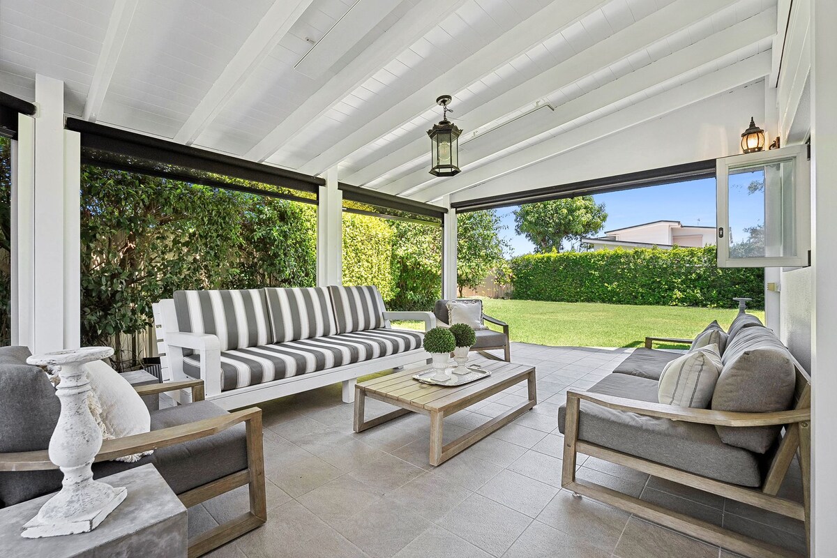 An outdoor living space is presented with a light-colored ceiling and spacious seating arrangements. A striped sofa and armchairs surround a wooden coffee table, topped with decorative items. Lush greenery is visible beyond the covered area, enhancing the sense of privacy and comfort.