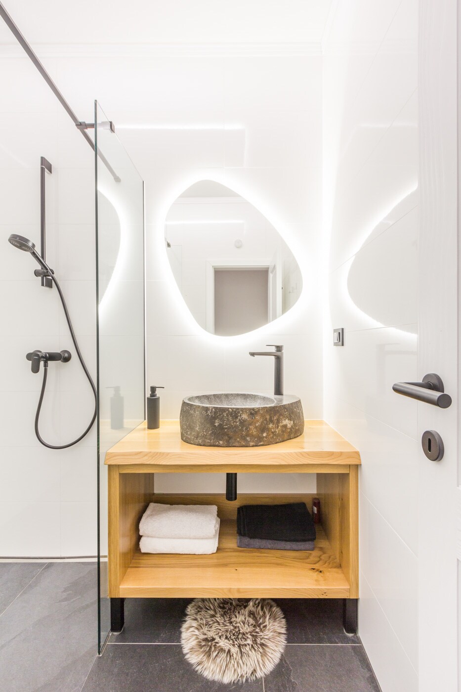 A modern bathroom features a sleek design with a stone wash basin set on a wooden vanity. Soft lighting accentuates the contours of the space, while a glass shower area is visible. Neatly arranged towels rest on the bottom shelf, enhancing the functional layout.