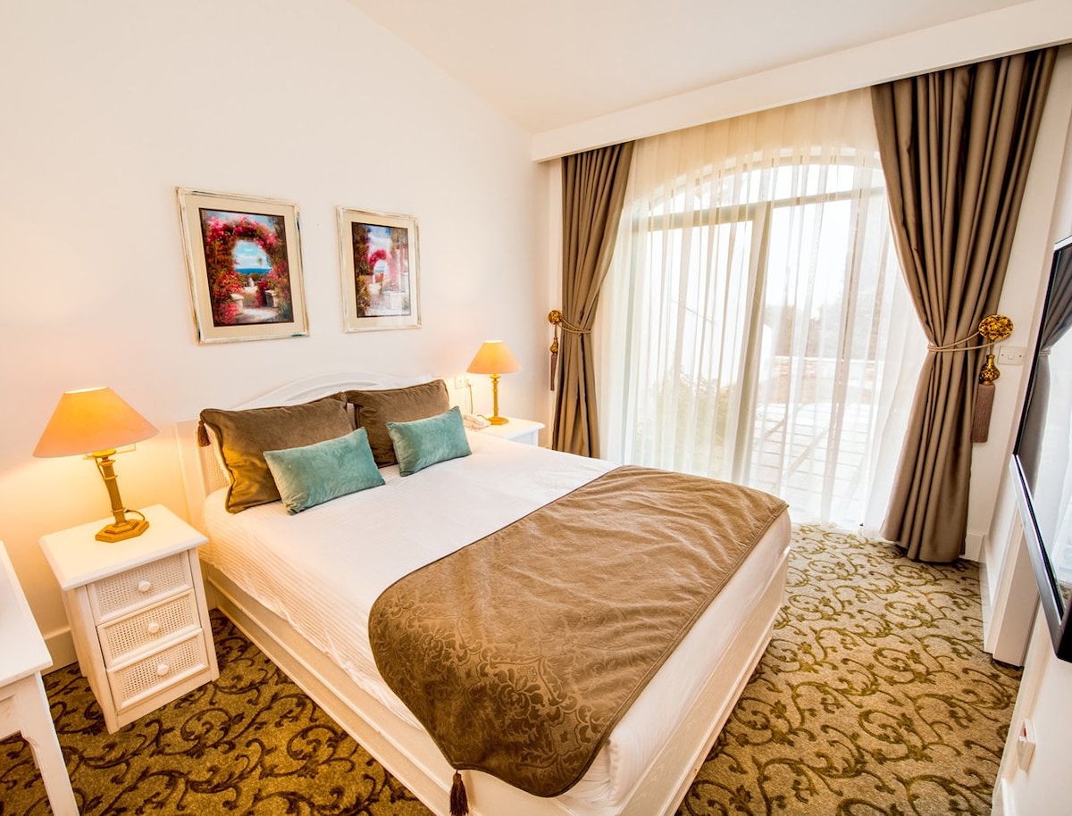 A well-lit bedroom featuring a spacious bed adorned with neutral linens and two decorative pillows. Bedside tables hold stylish lamps. Large windows with sheer curtains offer natural light and views of the outside. Framed artwork adds a touch of charm to the walls.