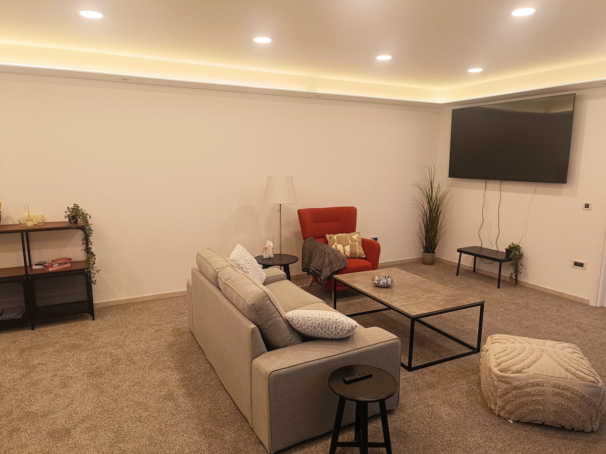 A spacious living area is presented, featuring a comfortable gray sofa and a vibrant red armchair. A large flat-screen television is mounted on the wall. The plush carpet adds warmth, while indirect lighting enhances the ambiance. A coffee table and side tables complete the inviting setup.