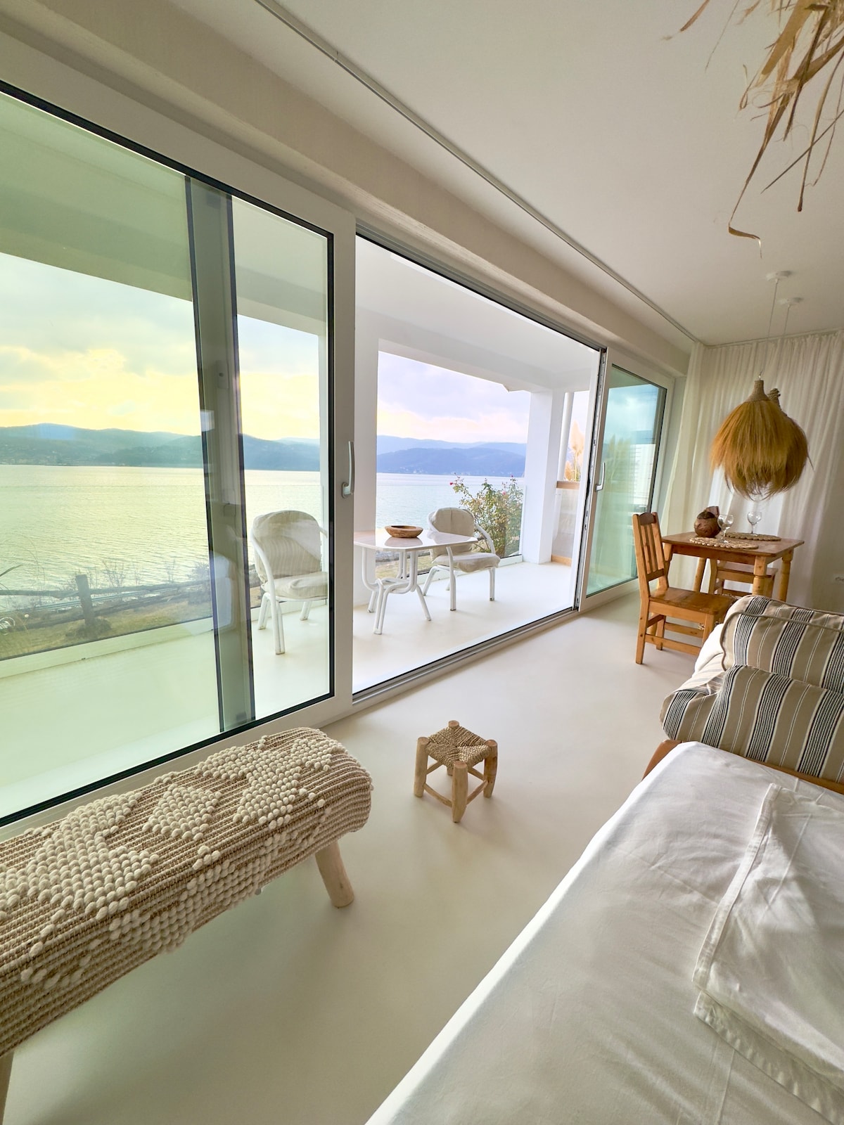 An open space living area offers a view through large glass doors that connect to a balcony overlooking the water. A dining table is set for four, while a cozy seating area includes a light-colored sofa and a woven stool, enhancing the serene atmosphere.