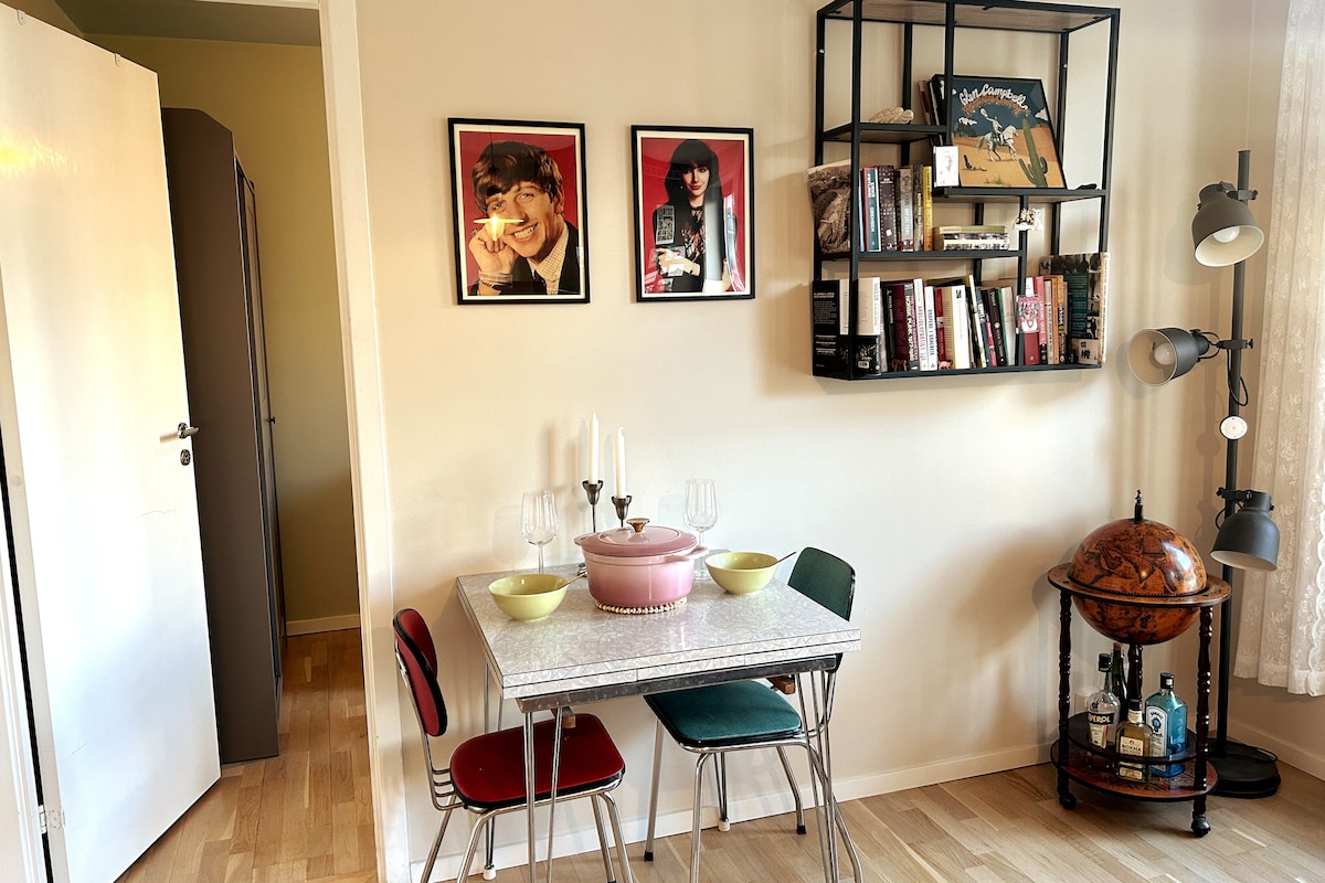 An intimate dining area is depicted with a small table surrounded by four mismatched chairs. A pink bowl is placed on the table, accompanied by two light-colored bowls. A wall-mounted shelf displays books and framed artwork, while a decorative globe adds charm to the space.