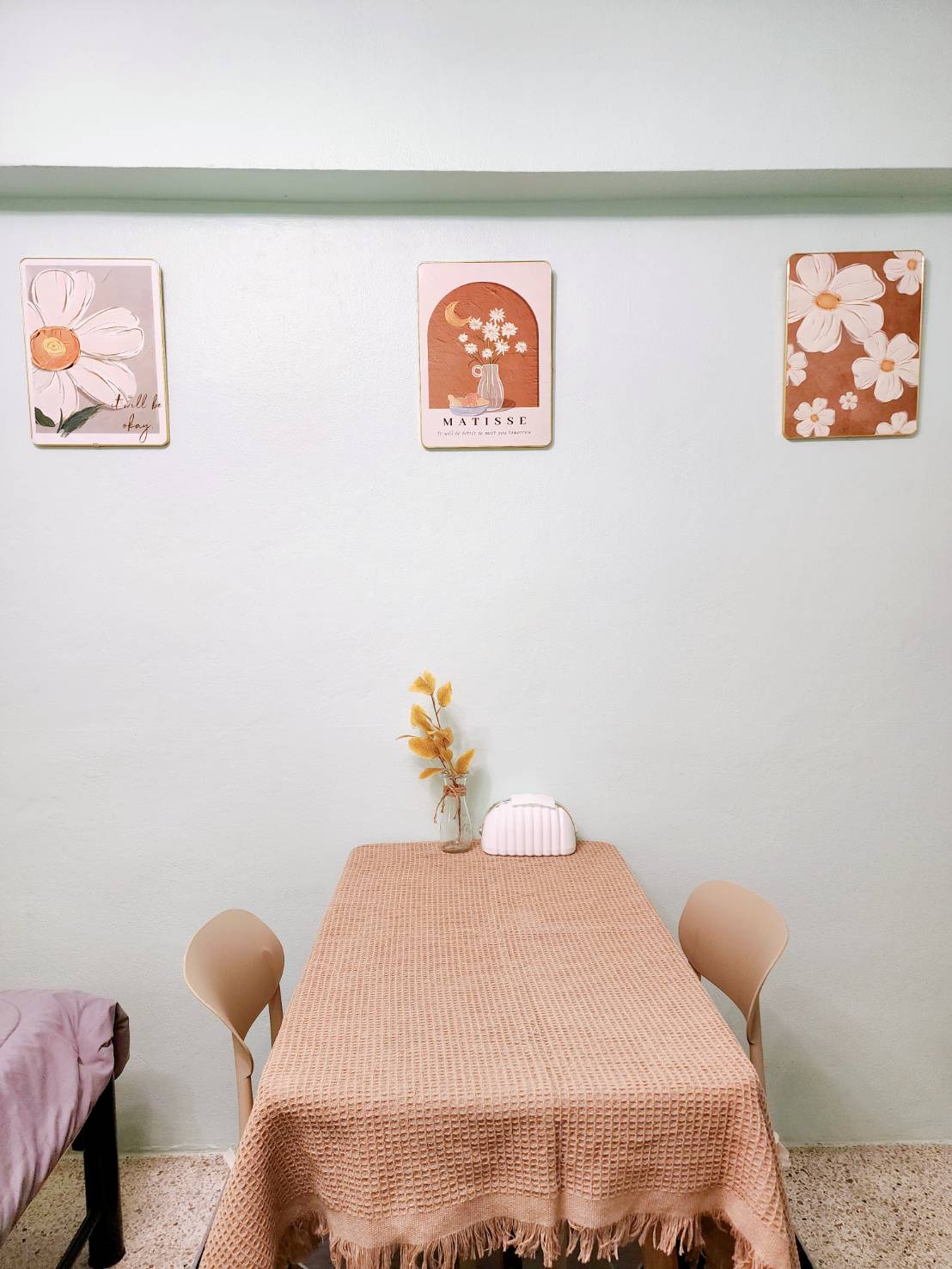 A small dining area features a simple table draped with a woven cloth and accompanied by two pastel-colored chairs. Above the table, three floral artworks are displayed against a soft green wall, while a vase with dried flowers adds a touch of decor.
