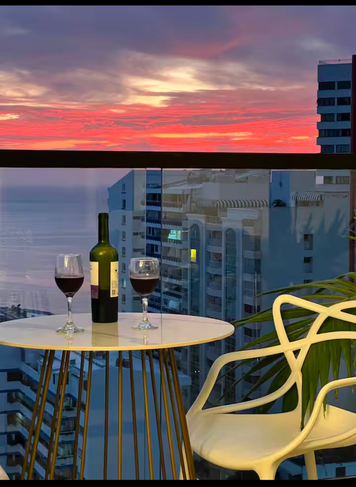 A scenic balcony is showcased with a small round table and two stylish chairs. Two glasses of red wine sit beside a bottle on the table, while a vibrant sunset reflects over the ocean. The view includes high-rise buildings in the background.