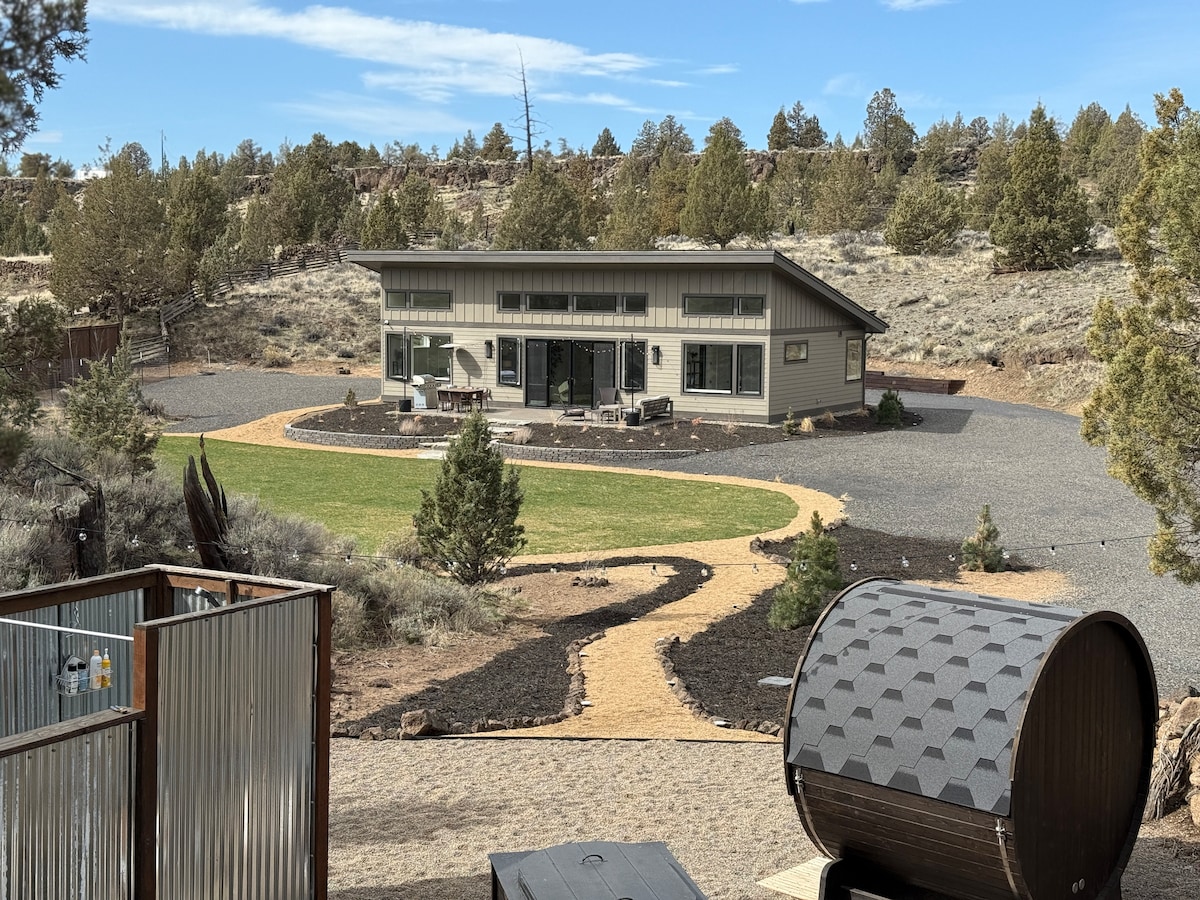 A modern two-bedroom home is situated on a landscaped expanse featuring a manicured lawn and gravel pathways. Surrounding terrain consists of native vegetation and panoramic views of the natural landscape. Outdoor seating areas are positioned on the patio, inviting relaxation in nature.