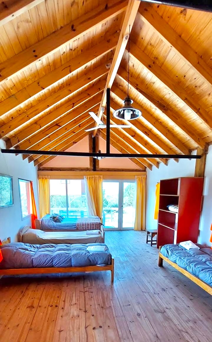 A spacious room features warm wooden beams across the high ceiling. Natural light floods in through glass doors that open to an outdoor space. Two beds are arranged on either side, complemented by bright orange curtains, while a simple bookshelf occupies one corner.