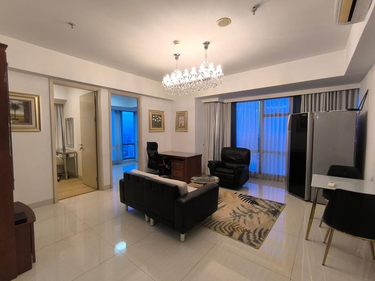A well-appointed living area is highlighted by a stylish chandelier and modern furnishings. A black leather chair and sofa are positioned on a rug, with a spacious desk nearby. Large windows bring in natural light, while a glimpse of the cityscape is visible through the glass.