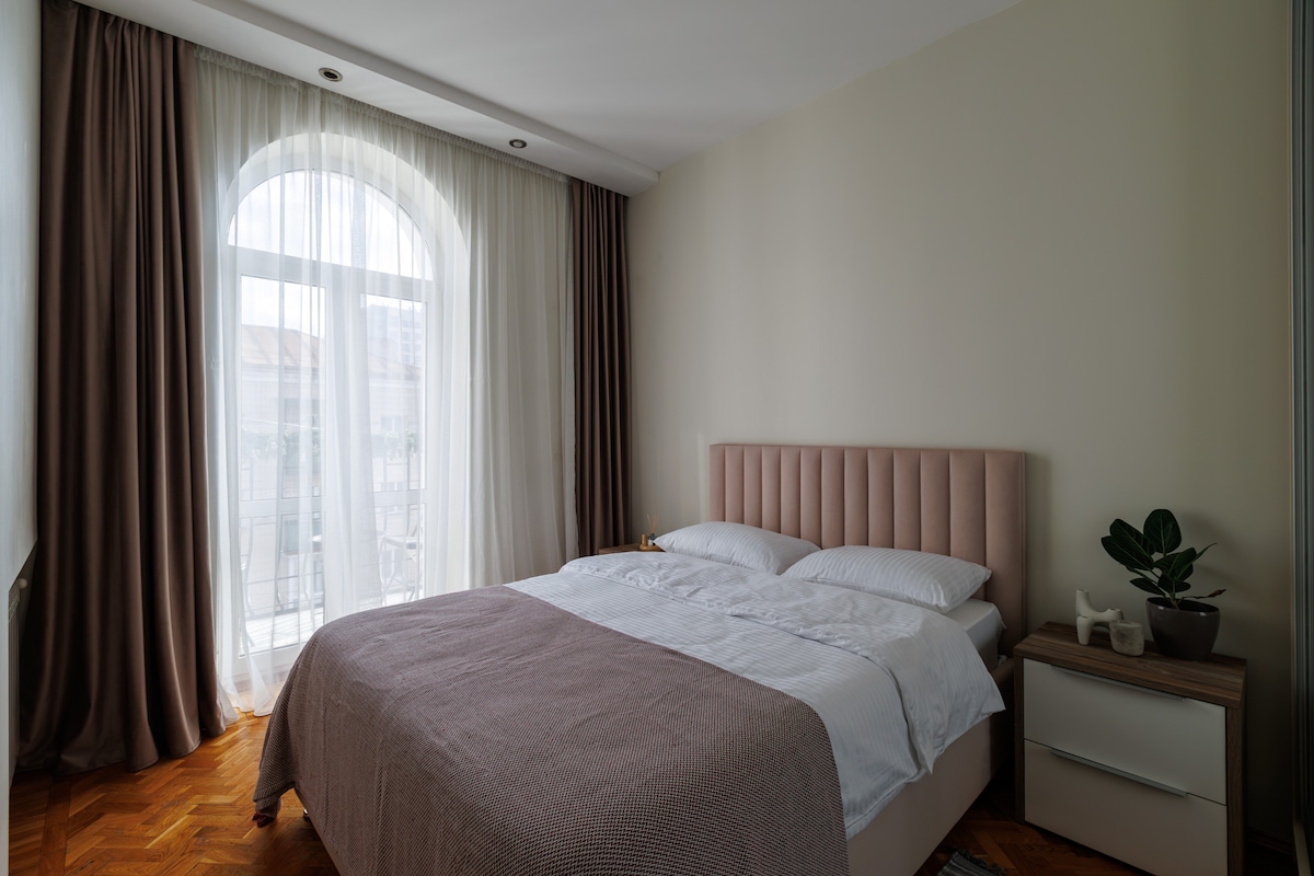 A serene bedroom features a spacious bed dressed in white linens and a light grey throw blanket. Natural light enters through an arched window, draped with sheer and dark curtains. A small plant is placed on a bedside table, accentuating the room's modern décor.
