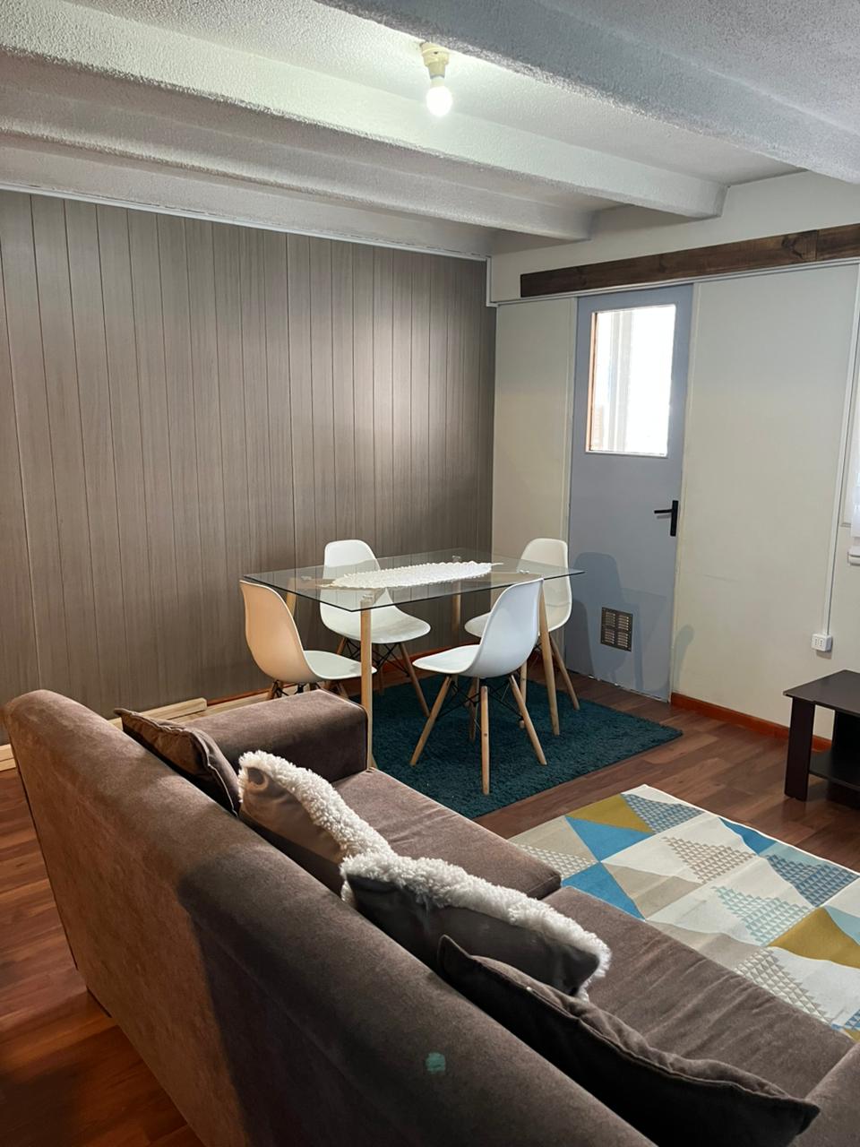 A dining area is positioned beside a window, featuring a glass table with four white chairs. A plush sofa in a neutral tone is placed nearby, complemented by a colorful area rug and a small dark coffee table, enhancing the room's cozy ambiance.