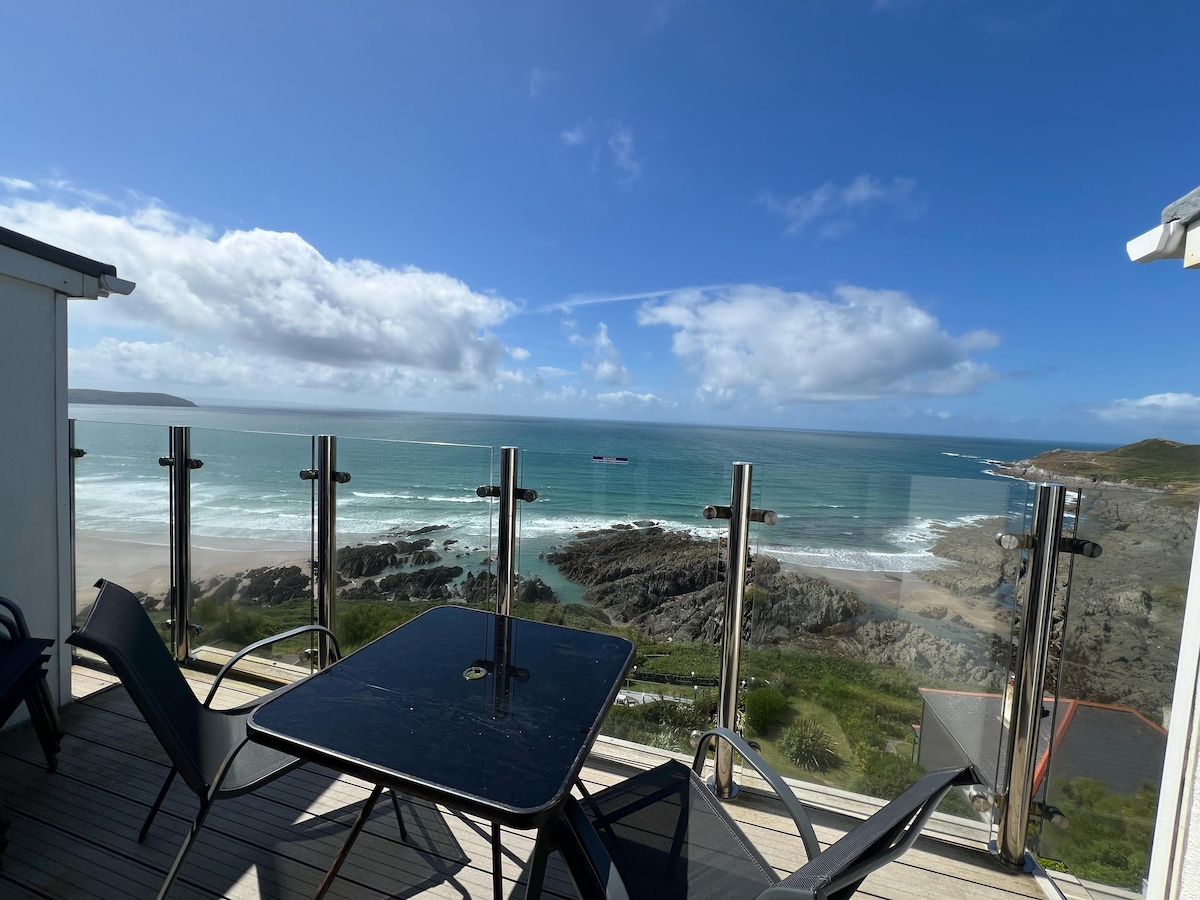 A decked patio area is visible, featuring black outdoor furniture with a table and chairs. The panoramic view captures the coastline, where the waves meet the sandy beach. A clear blue sky with scattered clouds complements the scenic beach landscape.