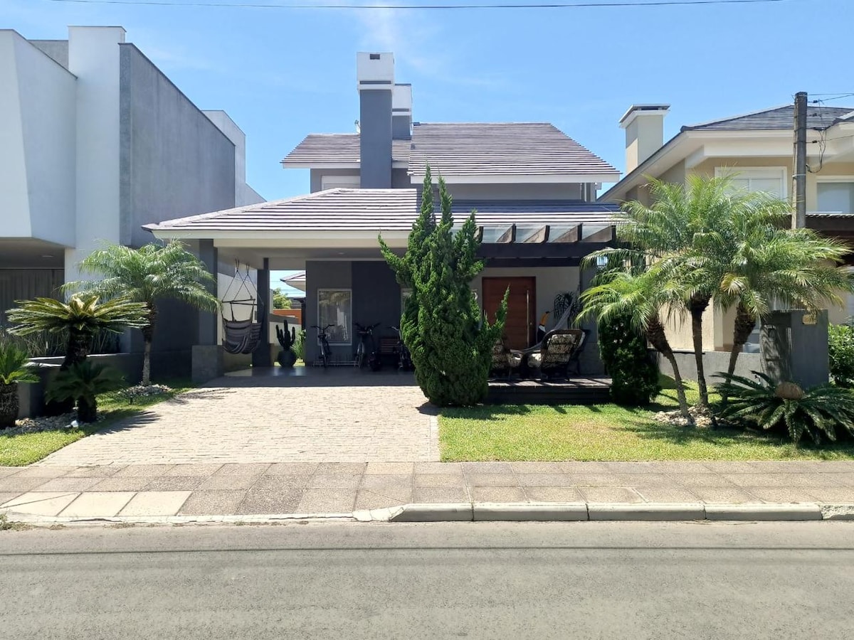 The exterior of a two-story house is presented, featuring a covered porch supported by columns. Lush greenery and palm trees adorn the front yard, while a paved pathway leads to the entrance. The roof is sloped, and sunlight bathes the façade in a warm glow.