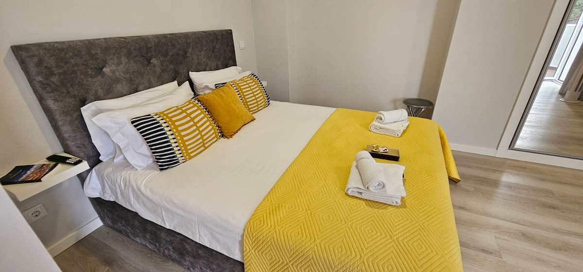A elegantly designed sleeping area features a plush gray headboard with a spacious bed dressed in white linens and a bright yellow throw. A neatly arranged set of towels is placed at the foot of the bed, with a mirror reflecting the soft tones of the room.
