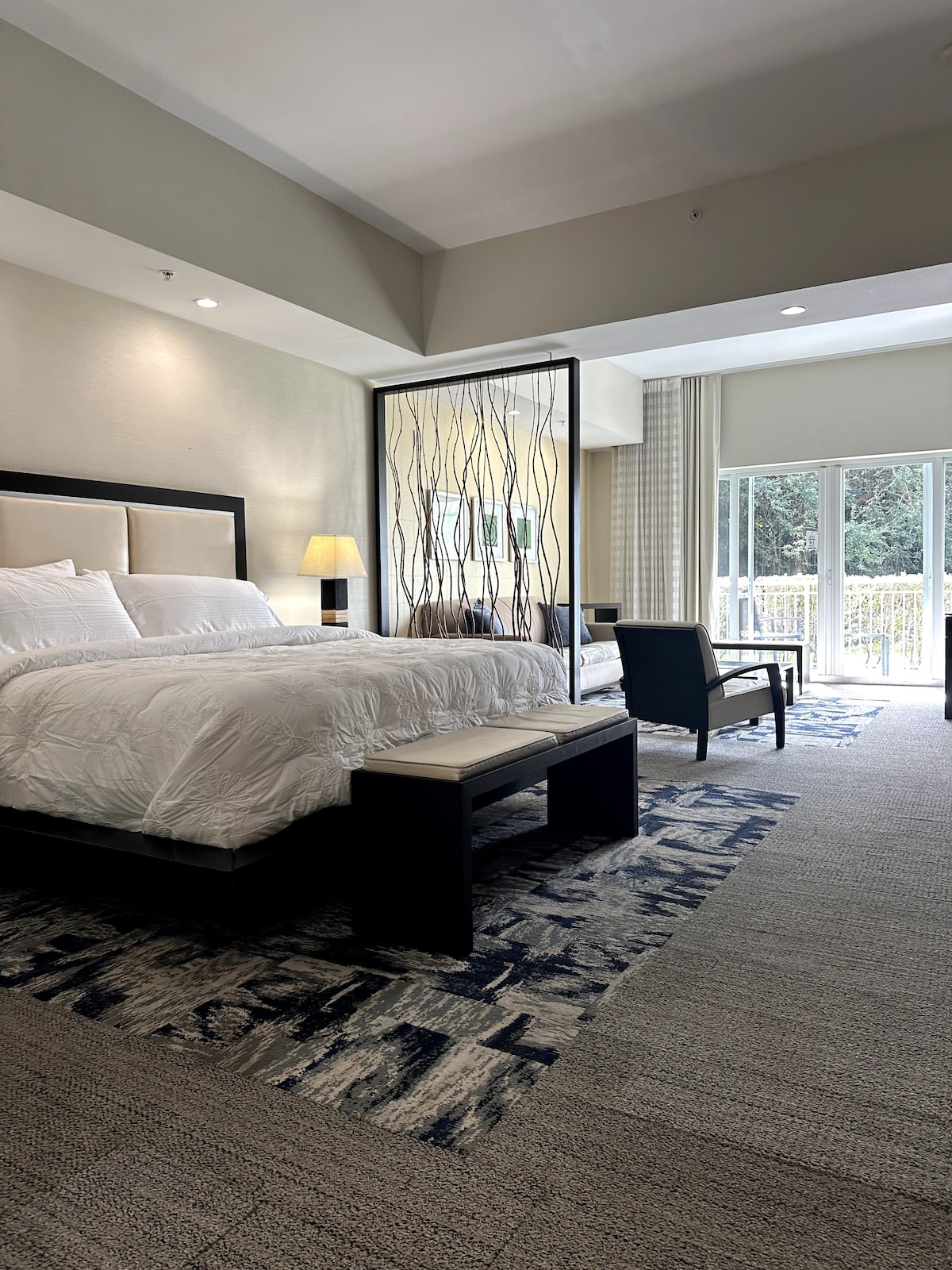 A spacious bedroom is highlighted by a king bed, separated from the seating area by a decorative room divider. Natural light enters through large glass doors, leading to a private balcony. A comfortable chair and simple bedside lamp complement the serene setting.