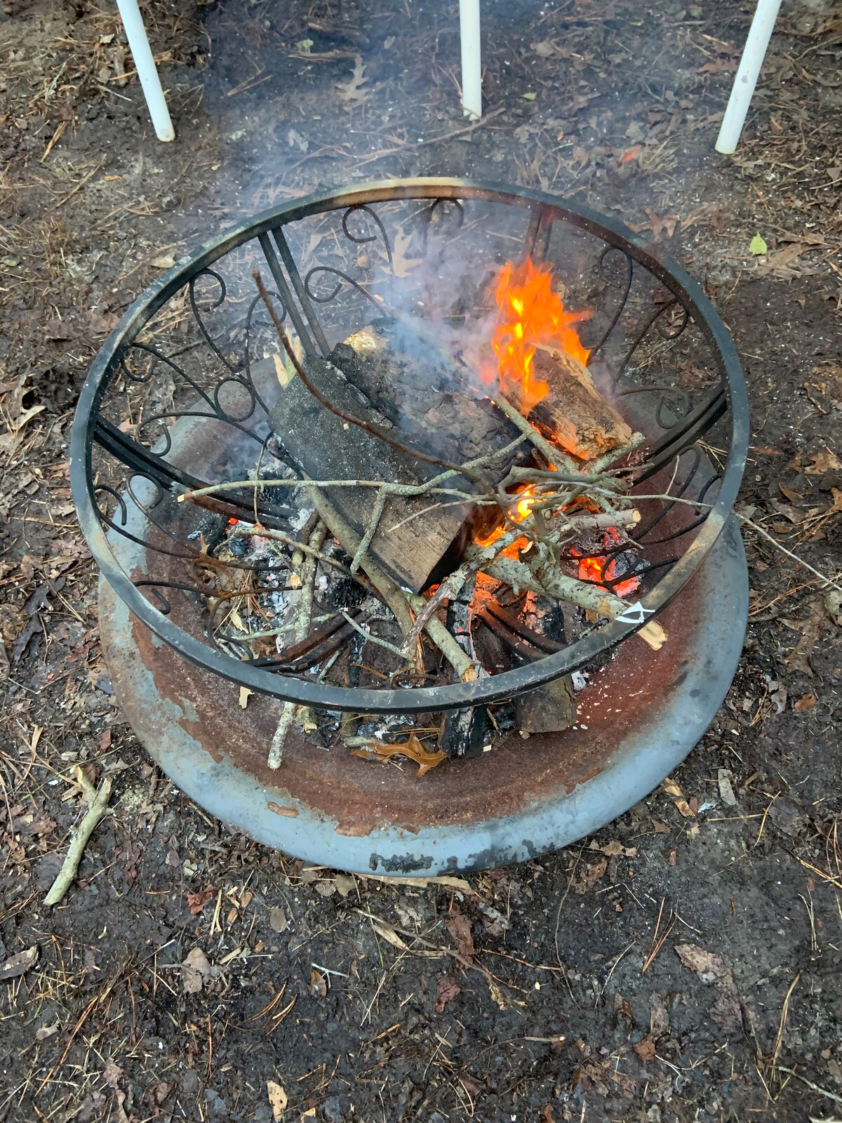 A circular fire pit is surrounded by a mix of stone and dirt ground, with flames brightly dancing over a bed of wood and kindling. A decorative metal ring encircles the fire, adding structure to the site for gathering and warmth.