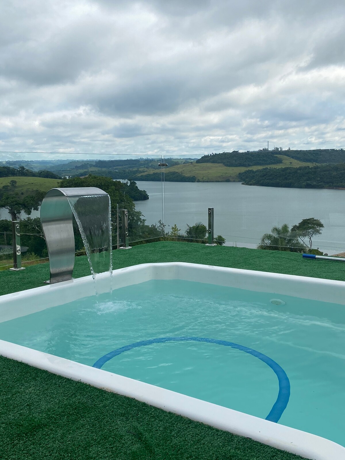 A sparkling pool is featured with a contemporary water feature, overlooking a serene lake and lush green hills in the background. The grassy area surrounding the pool adds a touch of nature, creating a relaxing spot ideal for leisure.