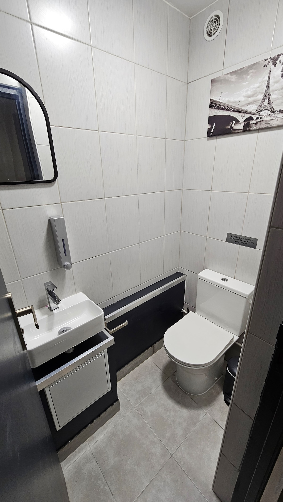 A bright bathroom is shown, featuring modern tiles and an elegant mirror. A compact sink and toilet are present, along with a framed image of the Eiffel Tower on the wall. Natural light enhances the clean, minimalistic design.
