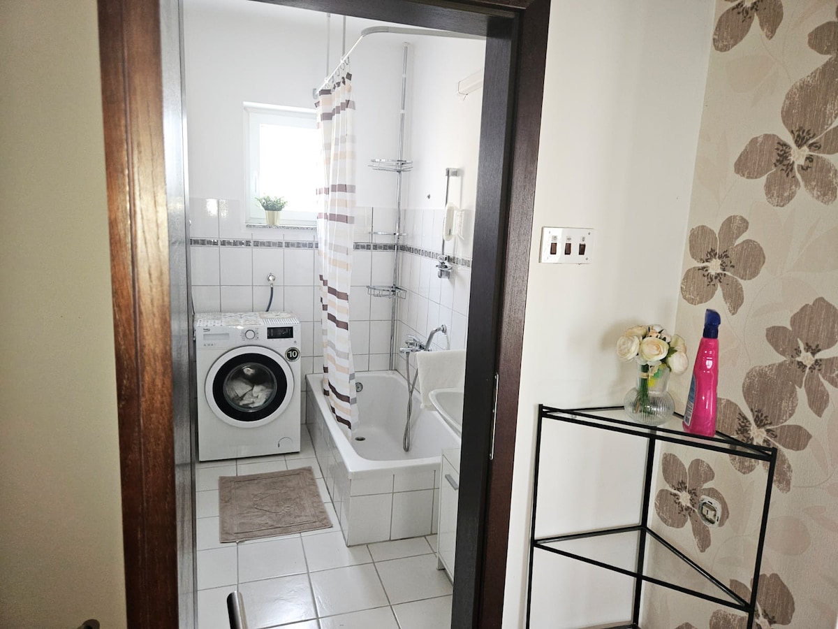 A bright bathroom is displayed featuring a bathtub with a shower, an enclosed washing machine, and shelving for toiletries. Light-colored tiles and a floral accent wall create a clean and calm ambiance. A small potted plant and flowers add a touch of freshness.