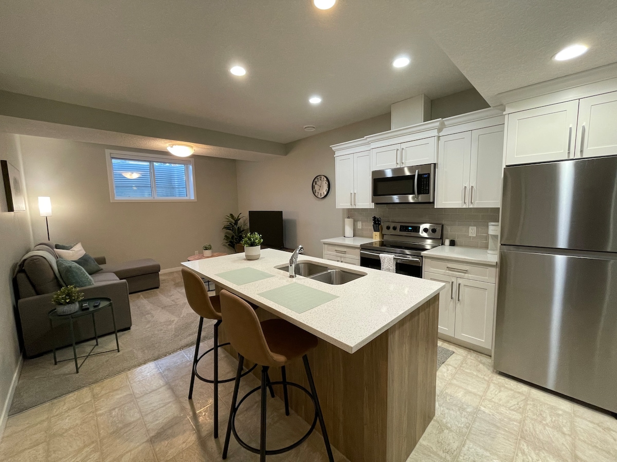 A modern kitchen area features a large island with sleek countertops and three bar stools. Stainless steel appliances are installed, including a refrigerator and oven. A cozy living space is visible in the background, complemented by natural light from a nearby window.
