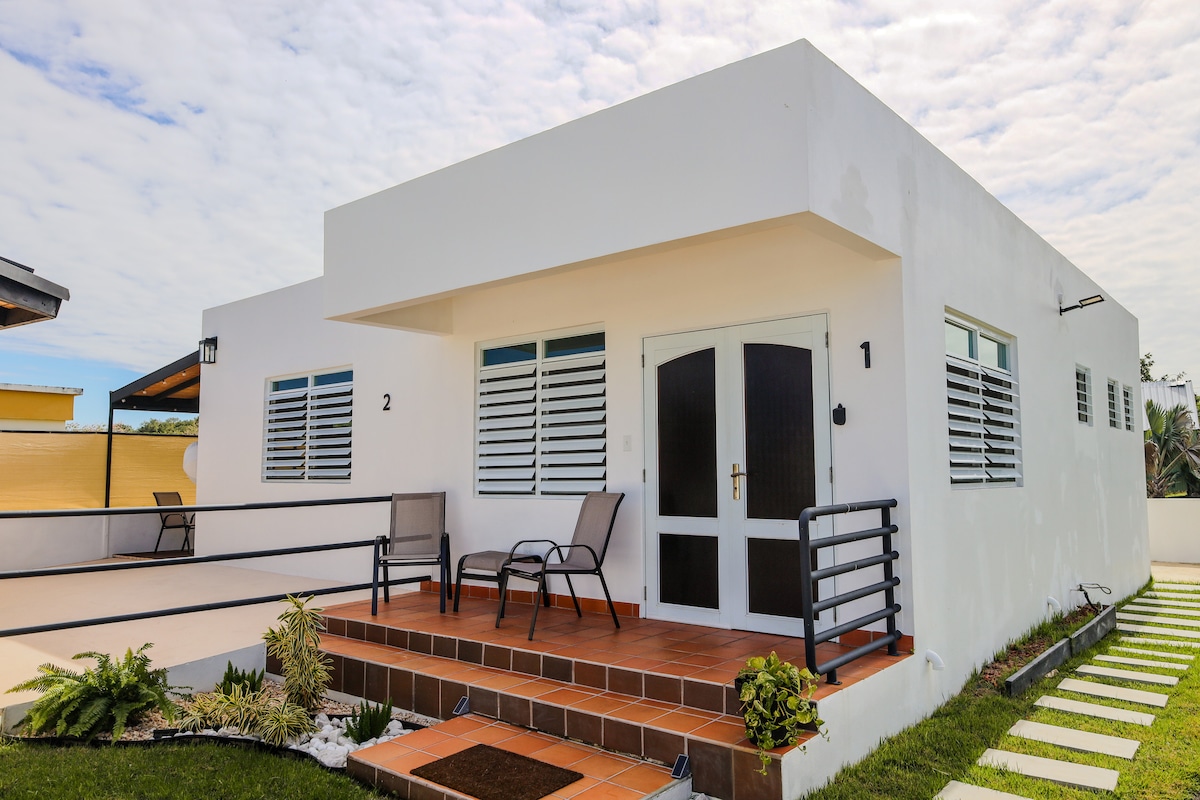 A modern white building features a welcoming entrance with two chairs positioned on a raised wooden deck. The façade showcases multiple windows with horizontal slats, allowing ample natural light. A neatly arranged pathway leads to the entrance, surrounded by landscaped greenery.