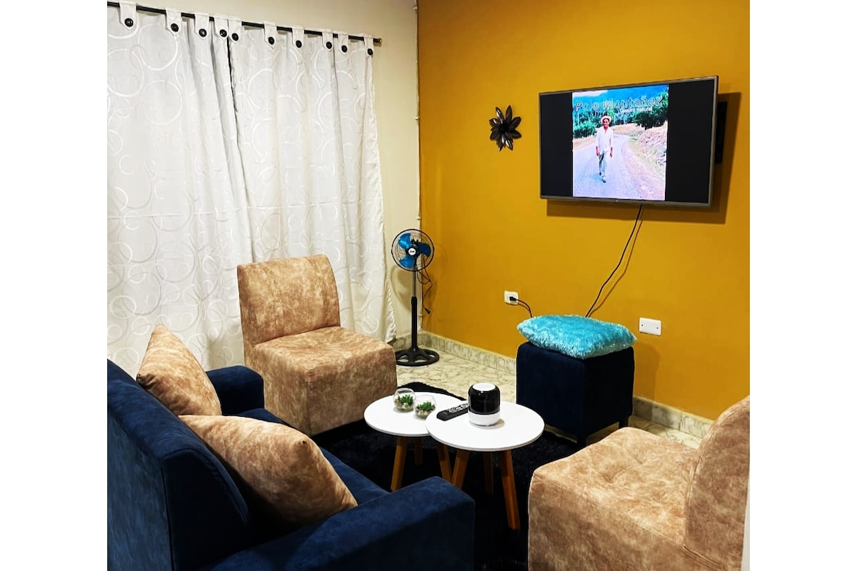 A cozy living area is presented with two soft brown armchairs and a blue sofa. A small round table holds decorative items, while a fan adds comfort. A flat-screen TV displays an outdoor scene against a backdrop of warm orange walls and sheer white curtains.