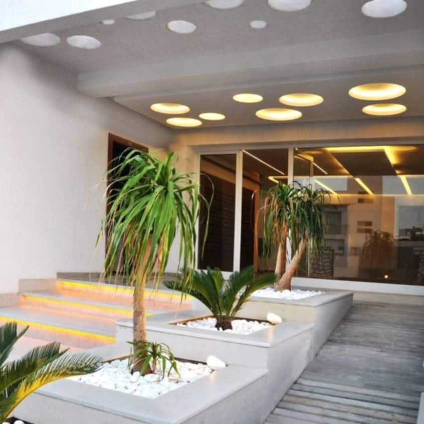 The entrance features a modern design with recessed circular lights illuminating the space. Potted palm plants are arranged in raised planters, surrounded by white stones. Steps lead towards large glass doors that provide a view into the elegantly lit interior.