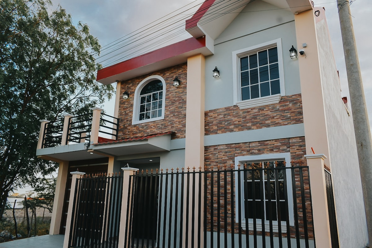 A modern two-story house is featured, showcasing a combination of brick and light-colored siding. The structure includes large windows, allowing natural light to brighten the interior. A decorative wrought iron fence surrounds the property, enhancing privacy and security.