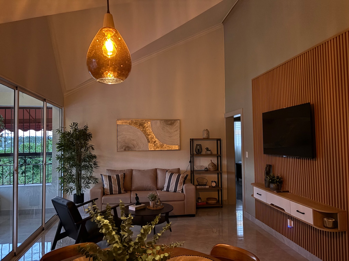 An inviting living area features a sectional sofa with neutral tones and decorative cushions, complemented by a coffee table. A tall plant adds greenery, while natural light pours in through large glass doors, revealing views of the outdoor scenery. A wall-mounted television is present, alongside decorative shelves.