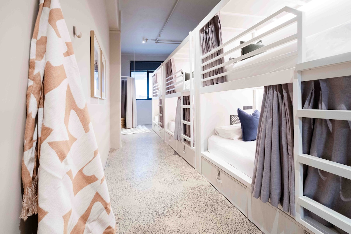 The dormitory features modern bunk beds with privacy curtains. A woven blanket hangs from a hook on the wall, adding a touch of comfort to the space. The polished concrete floor complements the minimalist decor, and natural light filters in through a window at the end of the hallway.