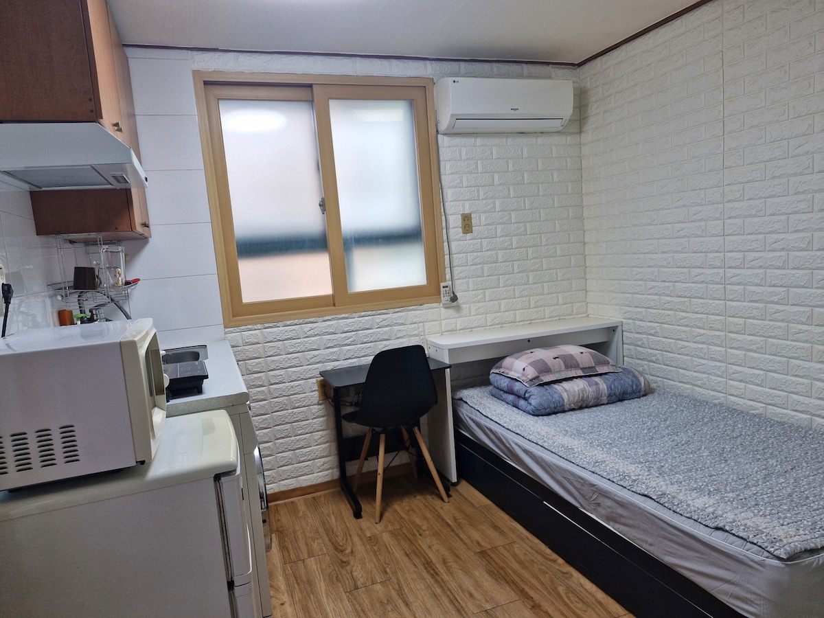 A cozy room is presented with a spacious bed and a simple desk with a black chair. The walls feature light-textured panels, and a window allows natural light to enter. A compact kitchen area is visible, equipped with a microwave and refrigerator.