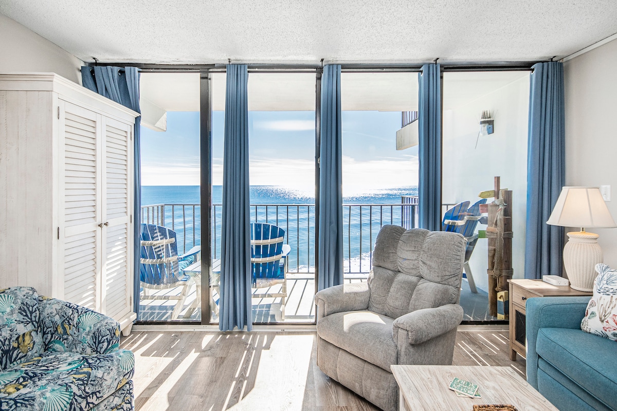 A sitting area features large glass doors framing an expansive ocean view. Soft blue drapes complement the light-colored furniture, including a plush recliner and a floral sofa. A small wooden table rests nearby. The sound of waves can be imagined from this inviting space.