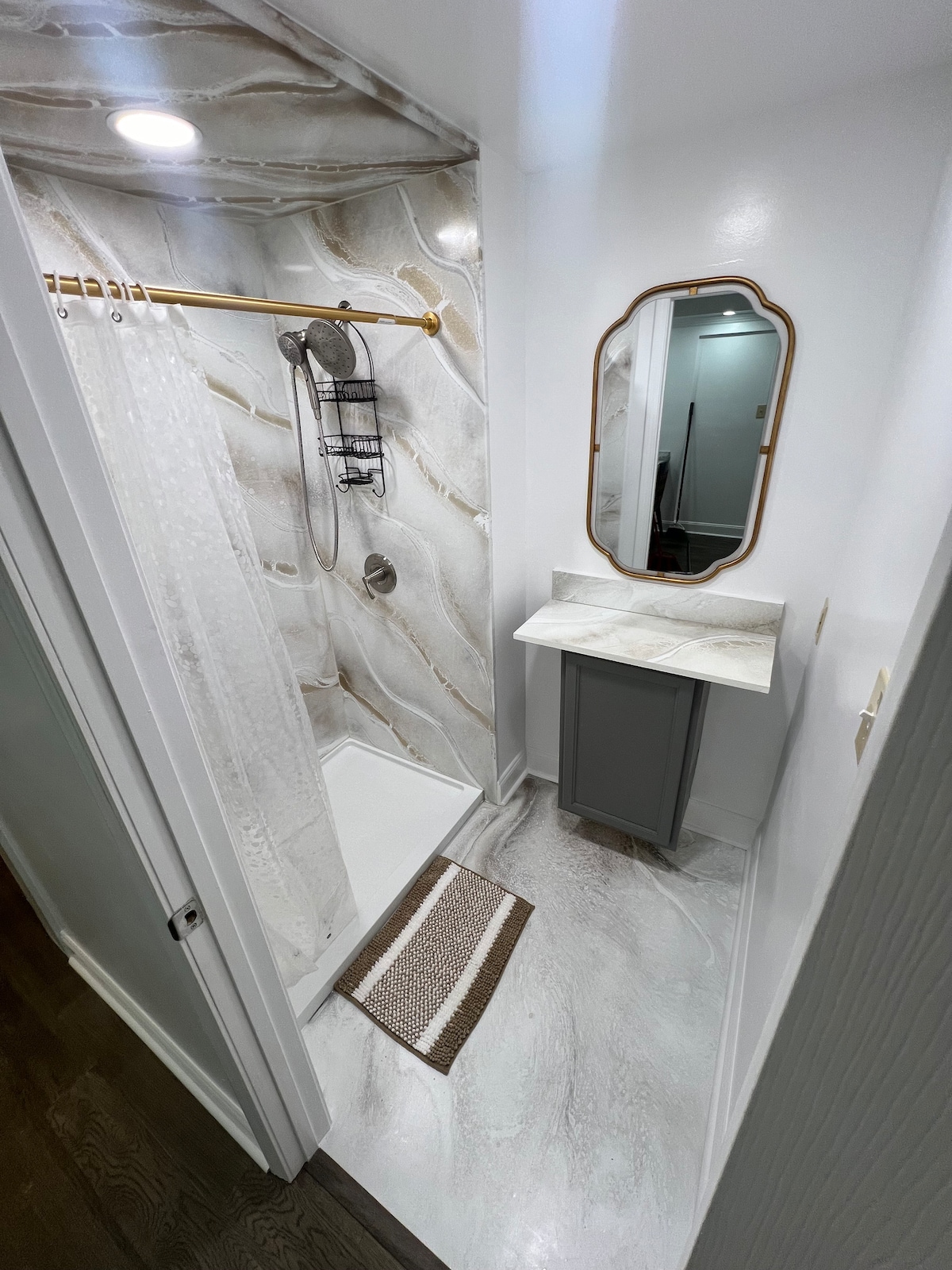 A walk-in shower is presented with a unique epoxy finish on the walls, highlighted by a modern showerhead and elegant fixtures. A small vanity with a marble-like surface and an oval mirror adds a touch of sophistication. The flooring displays a light, marbled design.