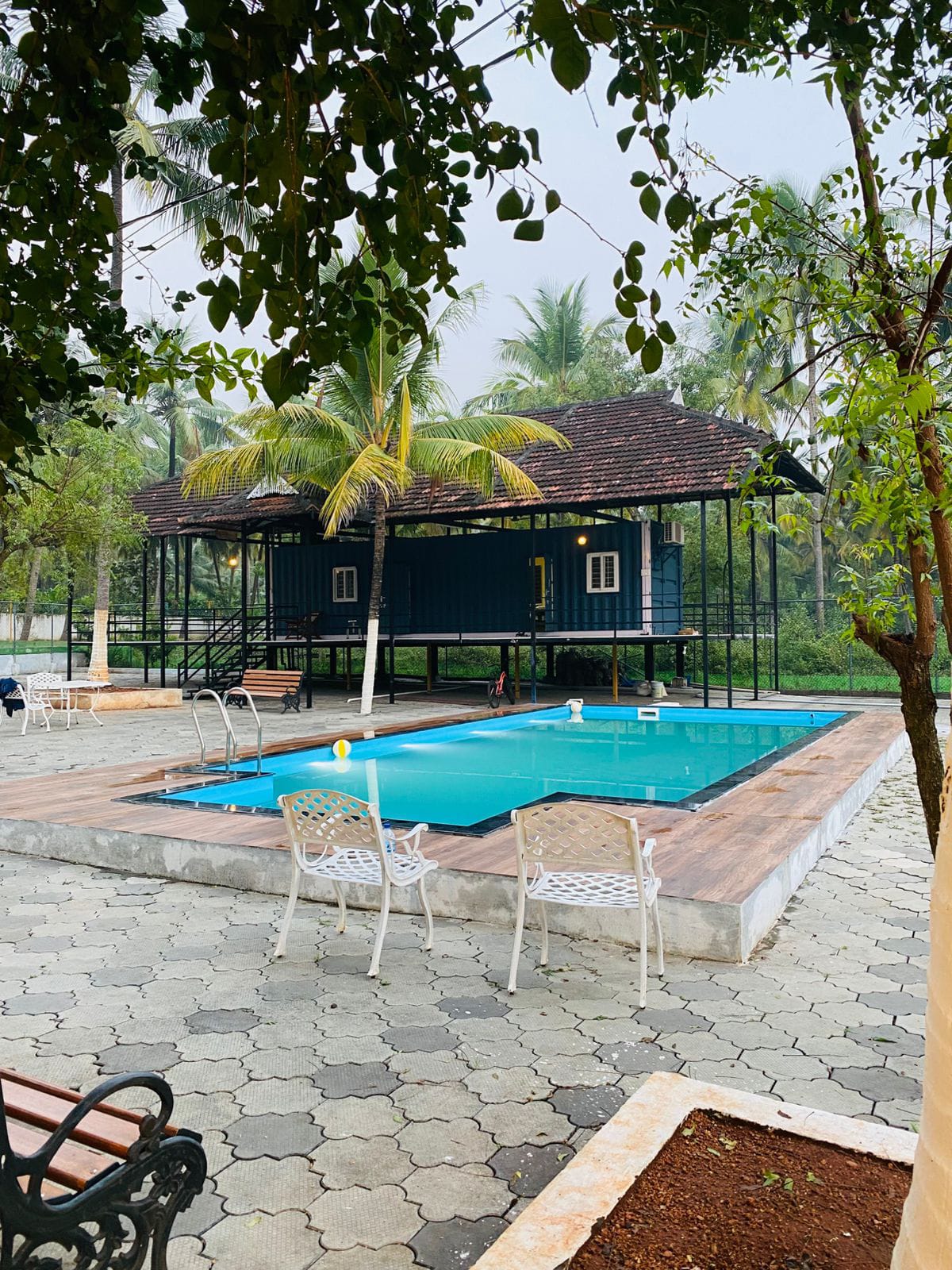 A serene poolside setting is depicted, featuring a clear blue swimming pool surrounded by smooth stone paving. A few white outdoor chairs are positioned nearby. Lush greenery and palm trees enhance the tranquil environment, with a stylish retreat structure visible in the background.