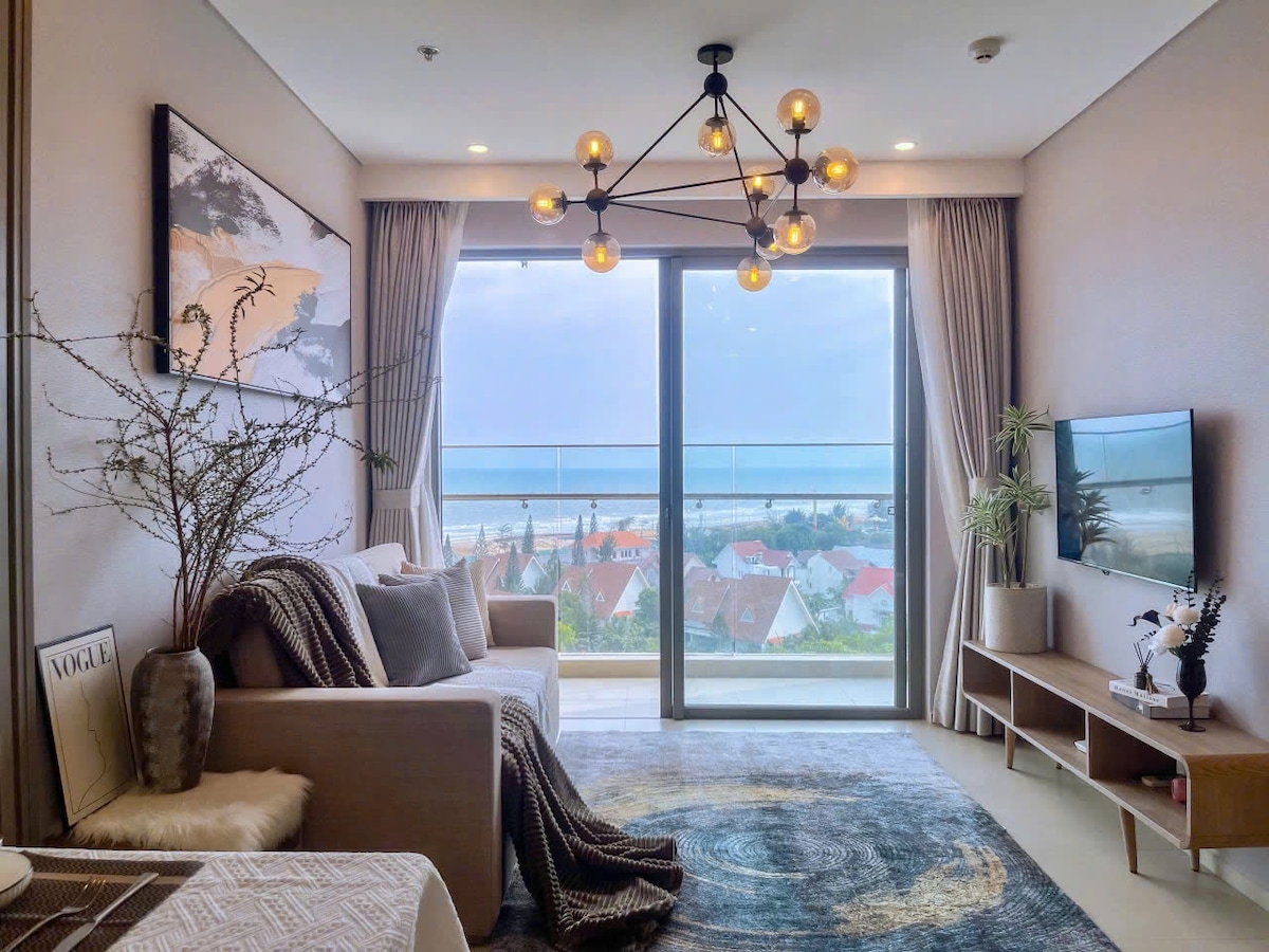 A bright living area features large glass sliding doors that offer expansive views of the ocean. Neutral-toned furnishings are complemented by a textured area rug and a modern light fixture. A small television and decorative elements on the media console enhance the space.
