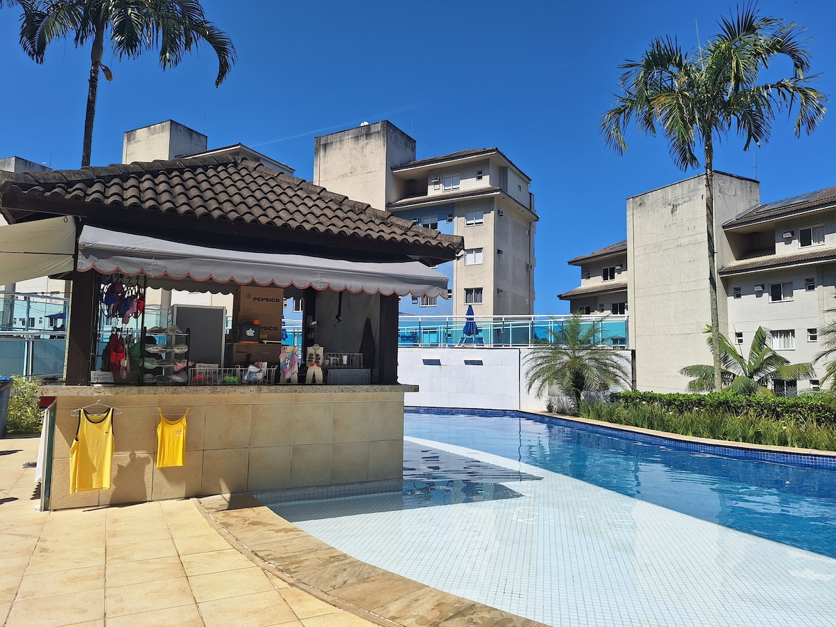 A spacious pool area is featured, with a snack bar under a tiled roof beside the water. Surrounding palm trees and modern buildings create a relaxed atmosphere, while the clear blue sky adds to the inviting scene.
