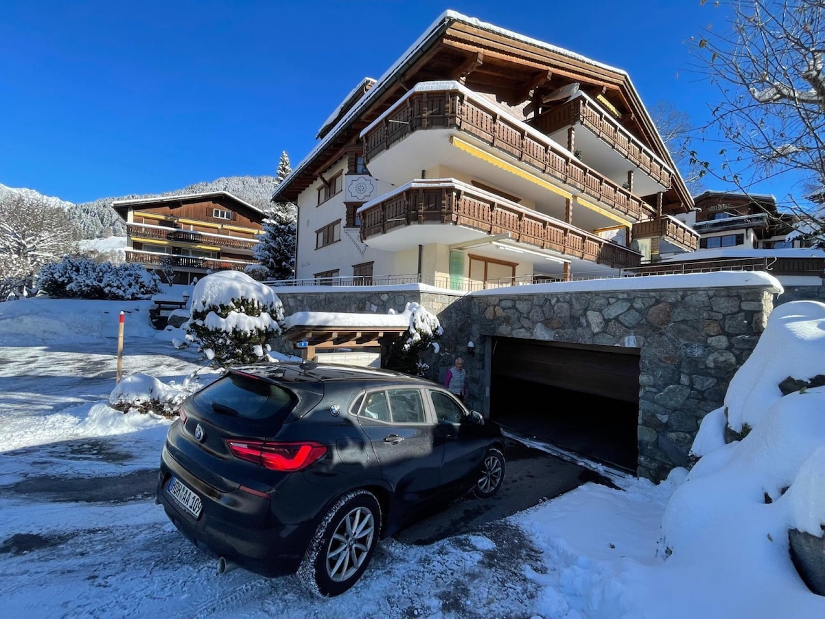 A modern apartment building is surrounded by snow-covered terrain, with wooden balconies and a stone exterior. A vehicle is parked in front of the entrance, leading to an underground garage. The backdrop features snow-capped mountains under a clear blue sky.