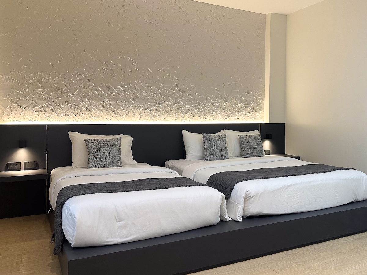 A well-appointed bedroom features two spacious beds, each topped with soft white linens and decorative pillows. Subtle lighting highlights a textured wall, creating a calming ambiance. Nightstands are positioned on either side, equipped with lamps for additional illumination.